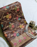 A folded brown silk saree with intricate floral motifs and a gold zari border, styled on a white fabric background alongside decorative jewelry.