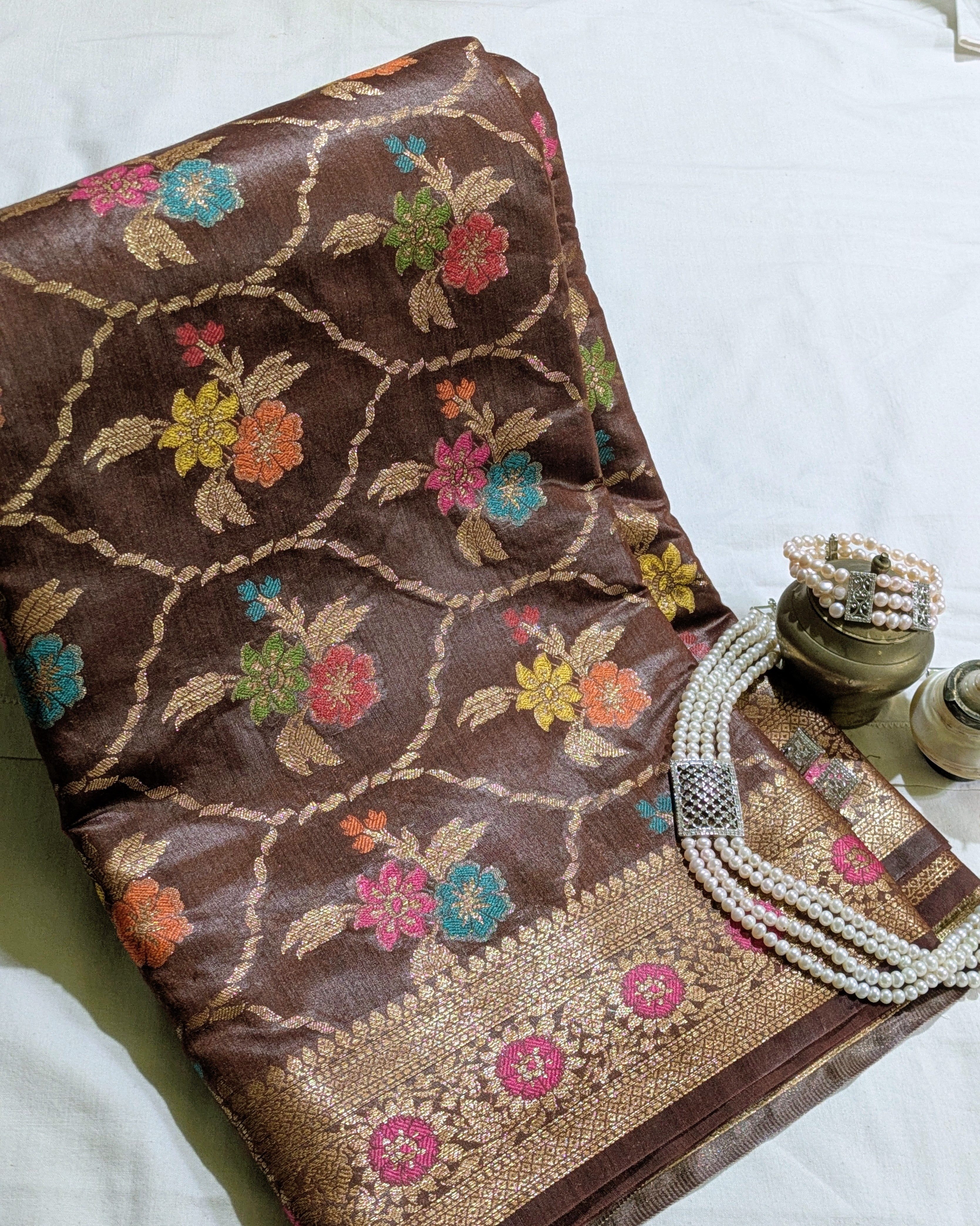 A folded brown silk saree with intricate floral motifs and a gold zari border, styled on a white fabric background alongside decorative jewelry.