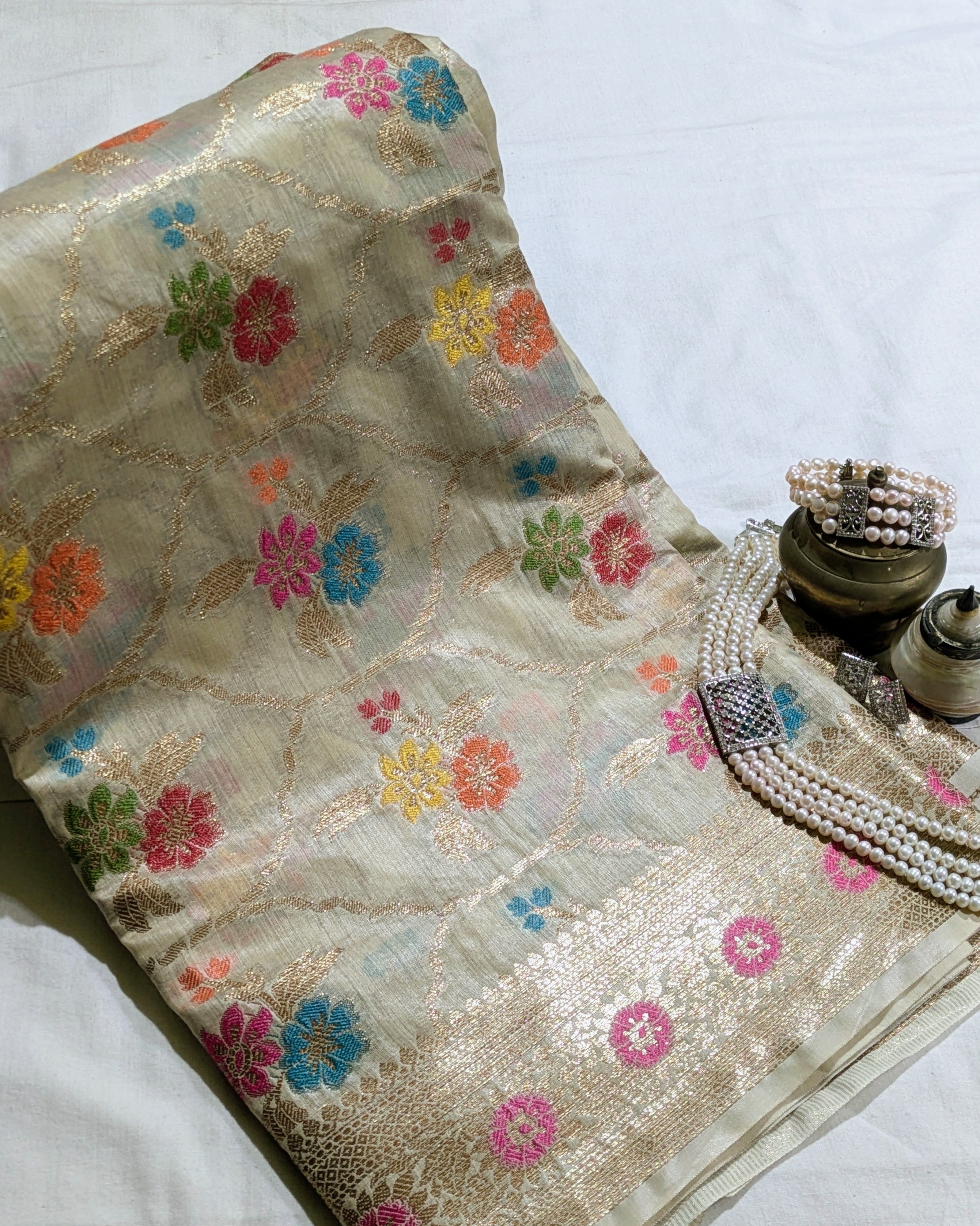 A beige silk saree with colorful floral motifs and a gold zari border, styled with pearl necklaces and brass weights on a white fabric background.