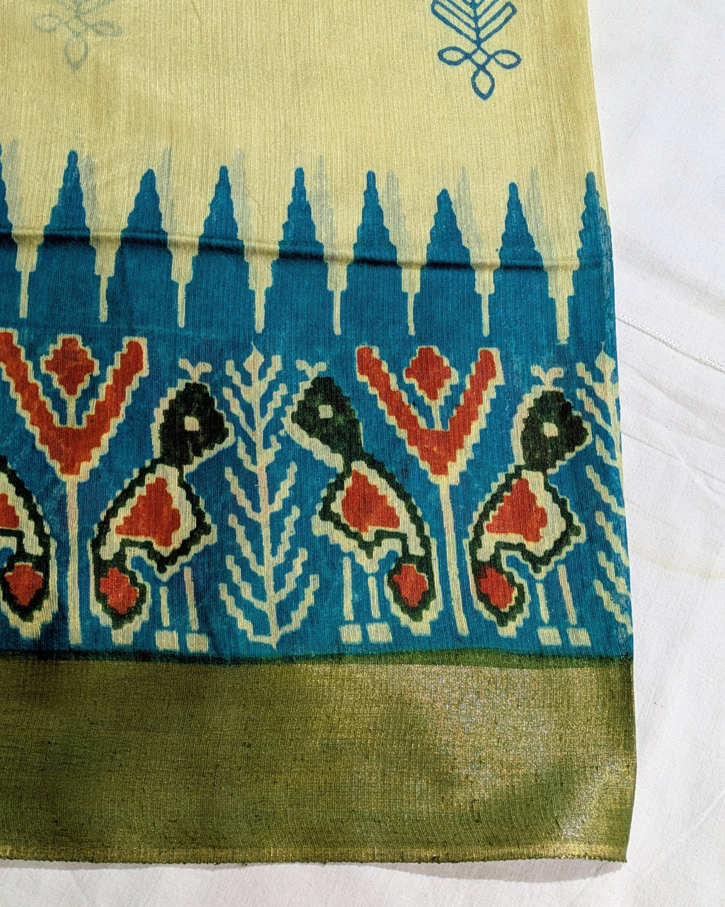 A close-up of a patterned fabric, likely a saree, featuring a cream-colored upper section with blue motifs, a wide blue border with red, black, and cream ikat-style fish and floral patterns, and a solid olive green border at the bottom.