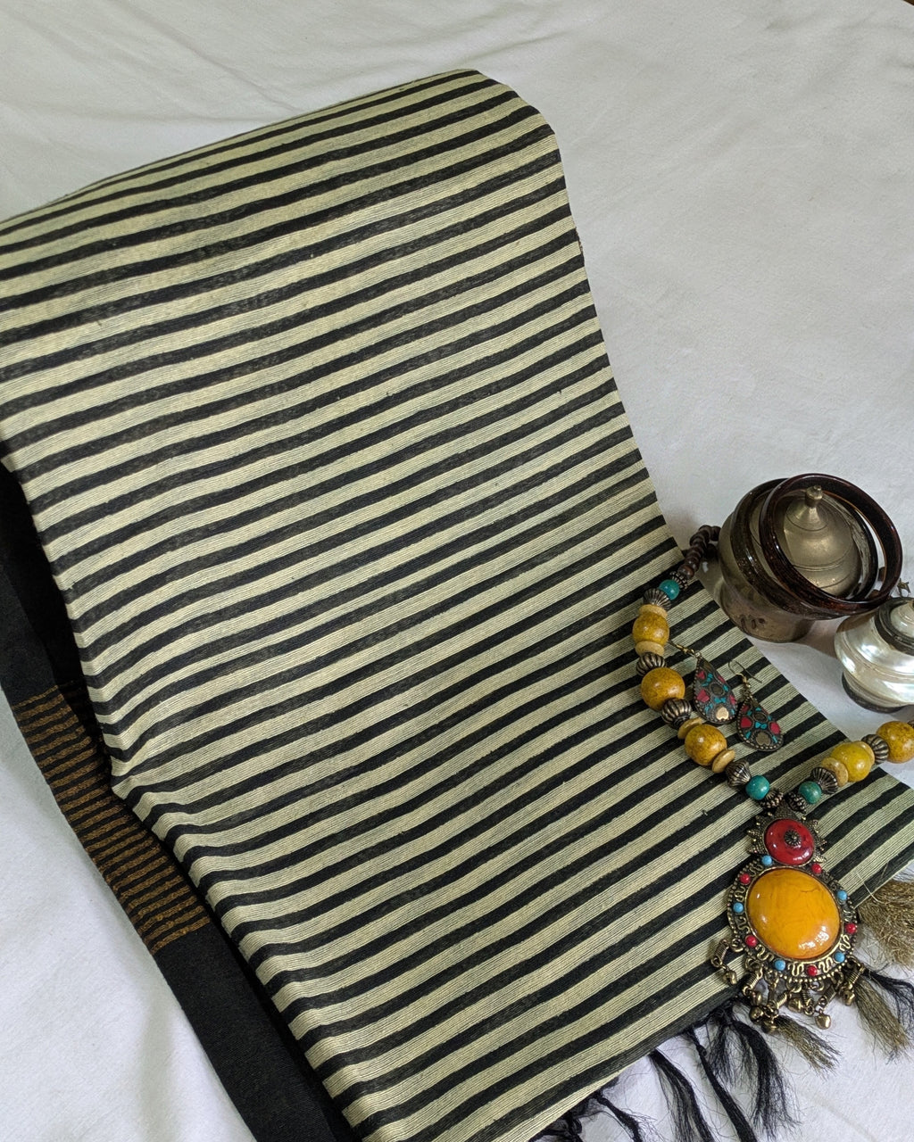 A folded striped textile, likely a scarf or shawl, in black and cream with fringe, styled on a white fabric background alongside decorative jewelry.