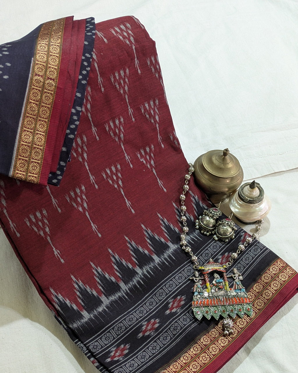 A folded traditional Indian saree or fabric piece in deep red with black and white ikat-style motifs and a navy border, styled on a white fabric background with decorative brass and silver containers and a beaded necklace.