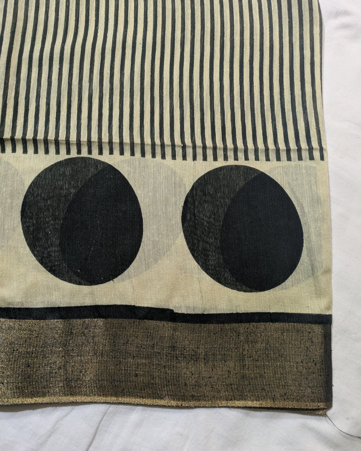 A close-up of a textile product, likely a scarf or sari, featuring a cream-colored base with black vertical stripes at the top, two large black oval motifs in the center, and a wide black border at the bottom.
