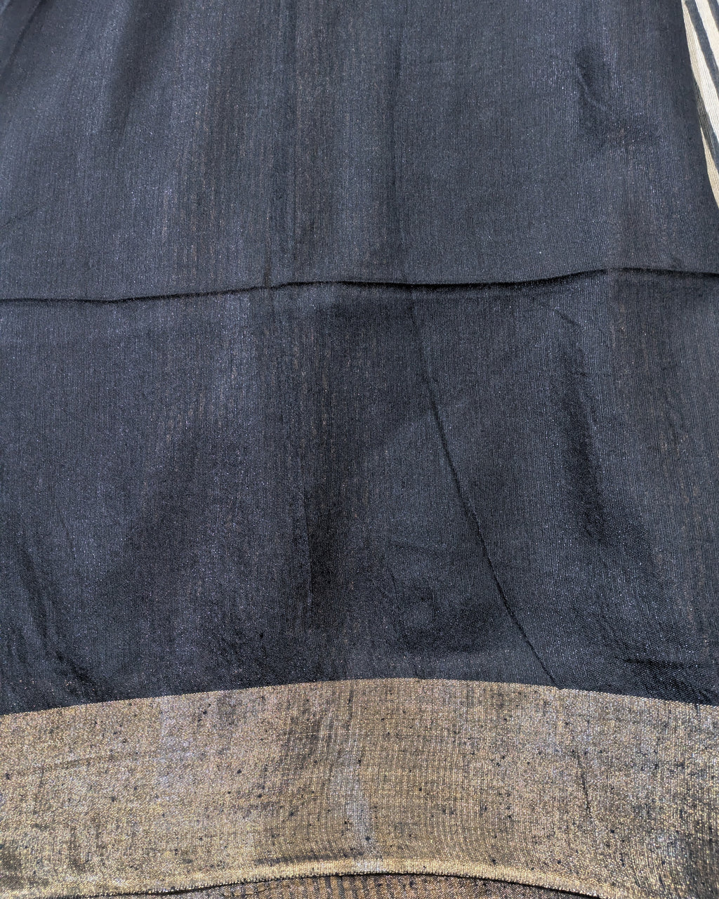 Close-up of a dark charcoal gray fabric with a textured, slightly coarse weave, featuring a horizontal seam and a contrasting metallic gold woven border at the bottom.
