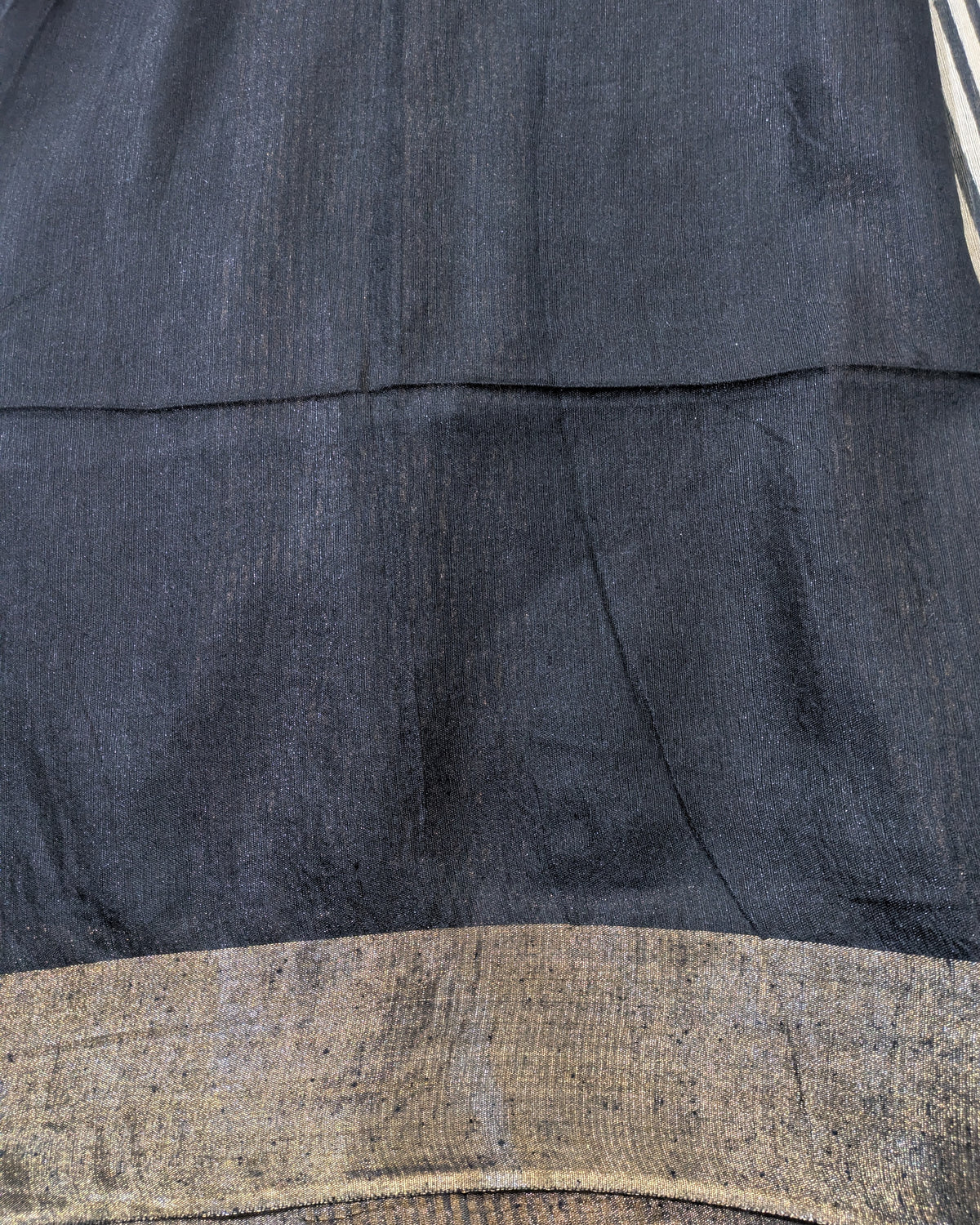Close-up of a dark charcoal gray fabric with a textured, slightly coarse weave, featuring a horizontal seam and a contrasting metallic gold woven border at the bottom.