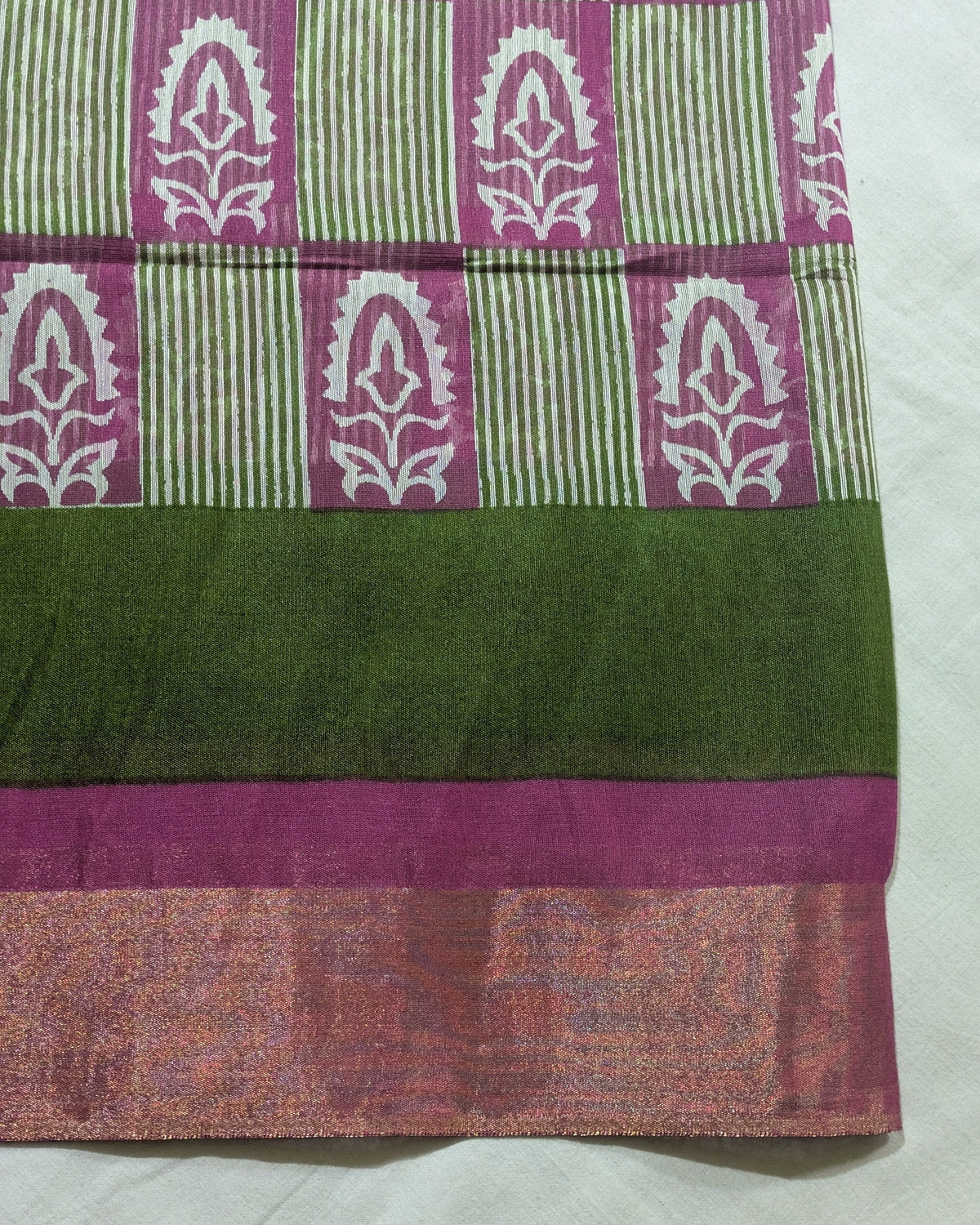A close-up of a patterned fabric, likely a saree or textile, featuring a magenta and green block-printed upper section with white motifs, a wide dark green middle band, and a metallic pinkish-gold border at the bottom.