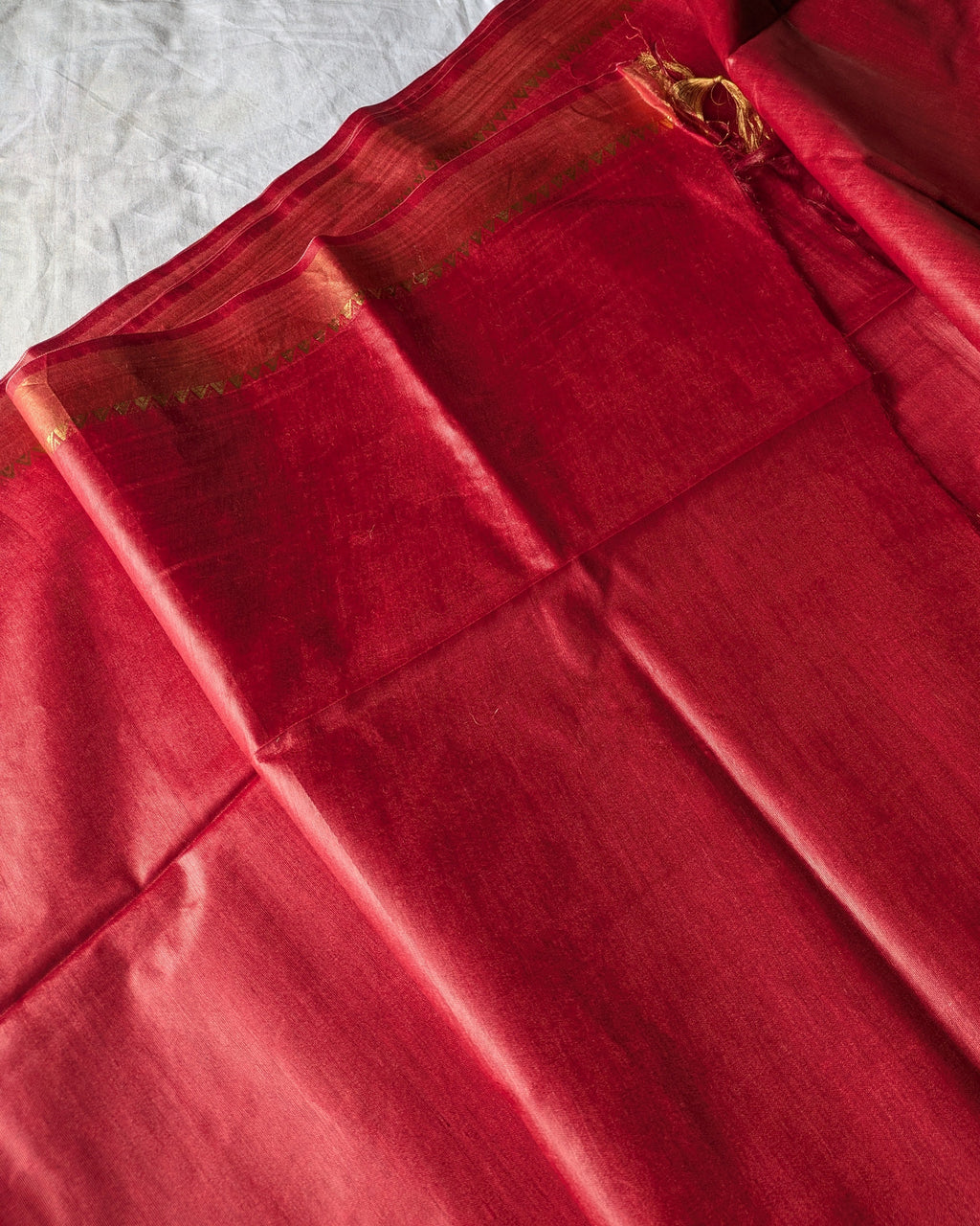 A close-up of a red silk saree fabric with a gold zari border, draped over a white fabric background.