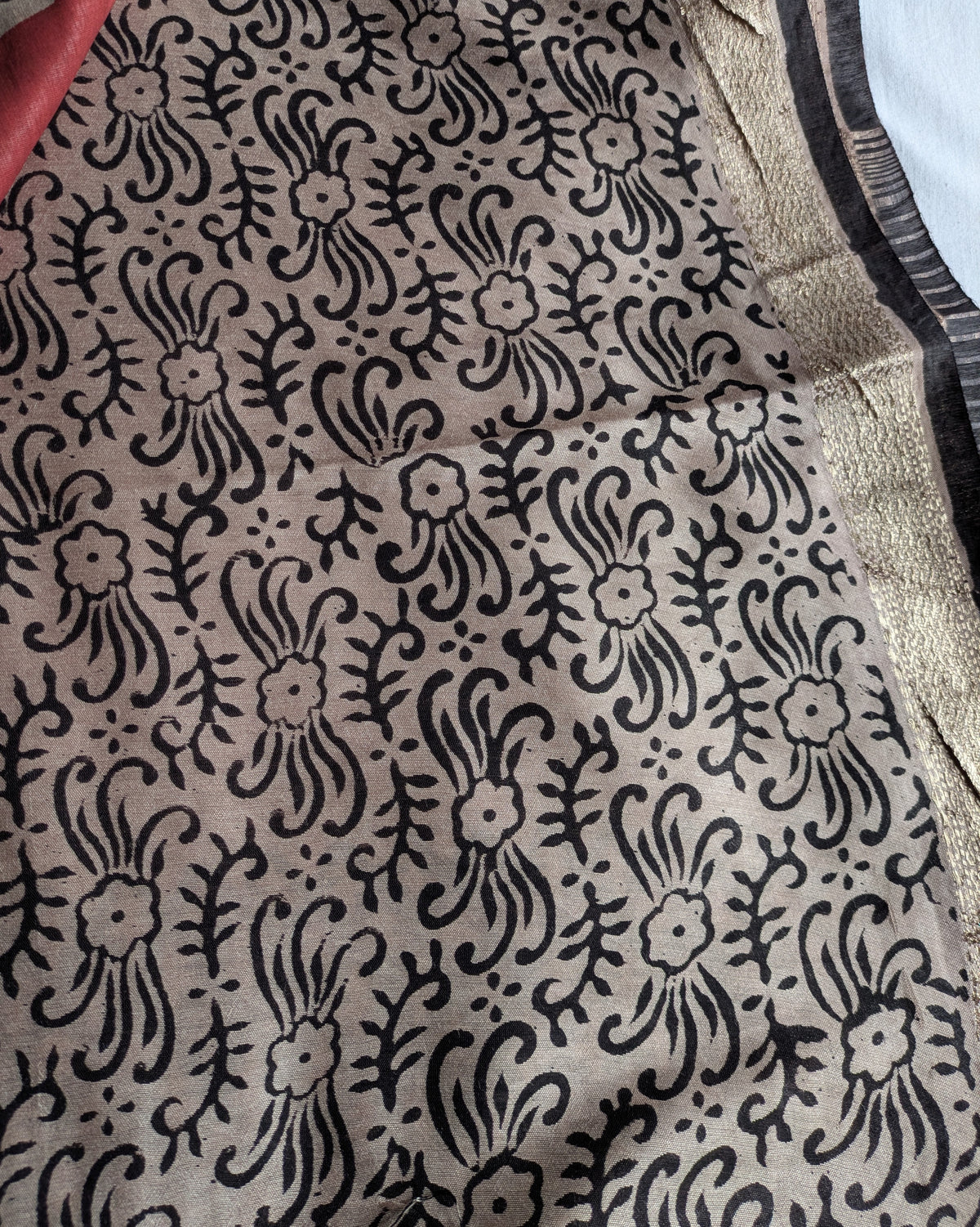 A close-up of a beige fabric with a black hand-block printed floral and paisley pattern, showing a metallic gold border on the right side.