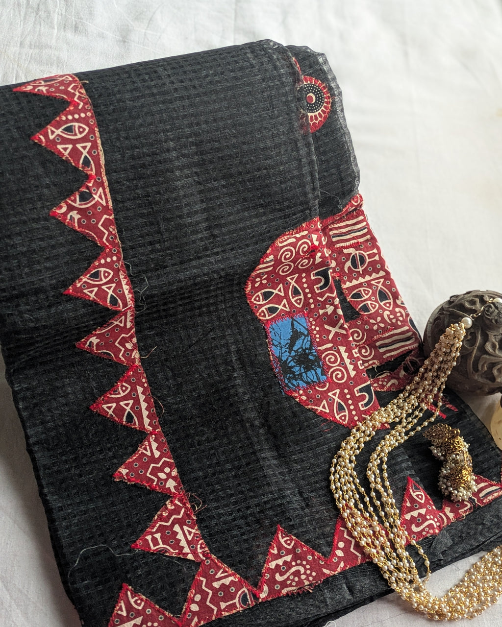 A folded black cotton fabric, likely a salwar kameez or sari, featuring red and beige geometric border patterns and a central red panel with a blue and black motif, styled with a gold beaded necklace on a white fabric background.