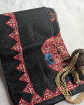 A folded black cotton fabric, likely a salwar kameez or sari, featuring red and beige geometric border patterns and a central red panel with a blue and black motif, styled with a gold beaded necklace on a white fabric background.