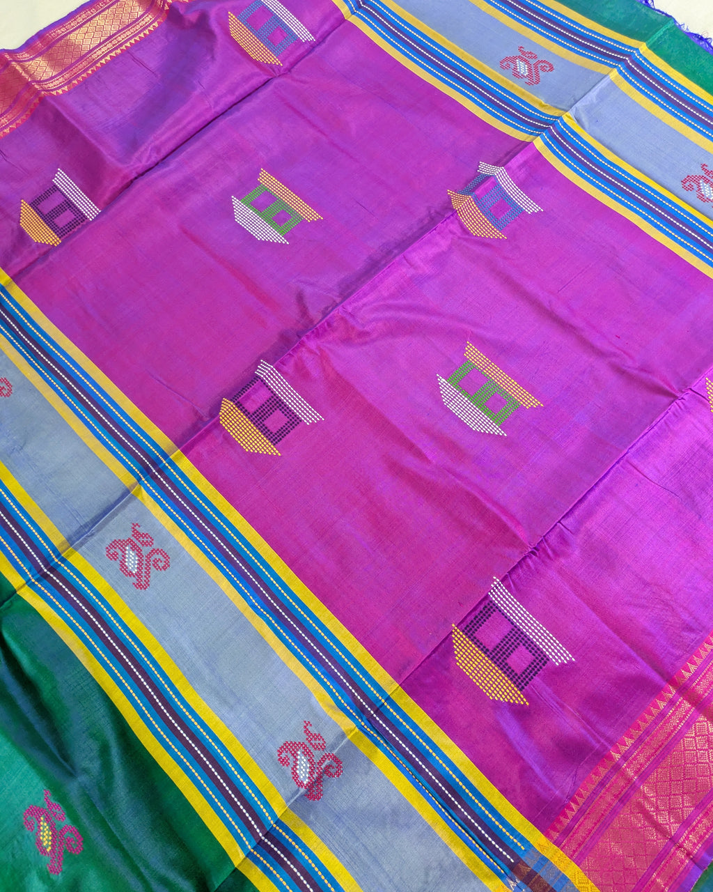 A close-up of a vibrant magenta purple silk saree body with woven geometric motifs, bordered by a light blue, yellow, and red striped pallu, and a green border visible in the bottom left corner.