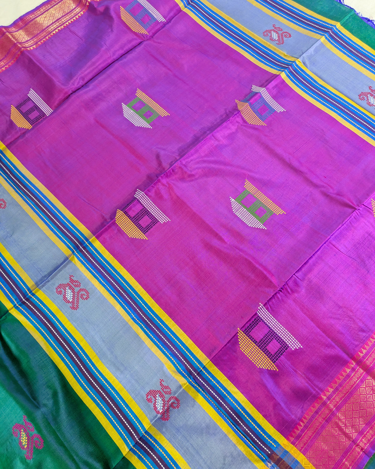 A close-up of a vibrant magenta purple silk saree body with woven geometric motifs, bordered by a light blue, yellow, and red striped pallu, and a green border visible in the bottom left corner.