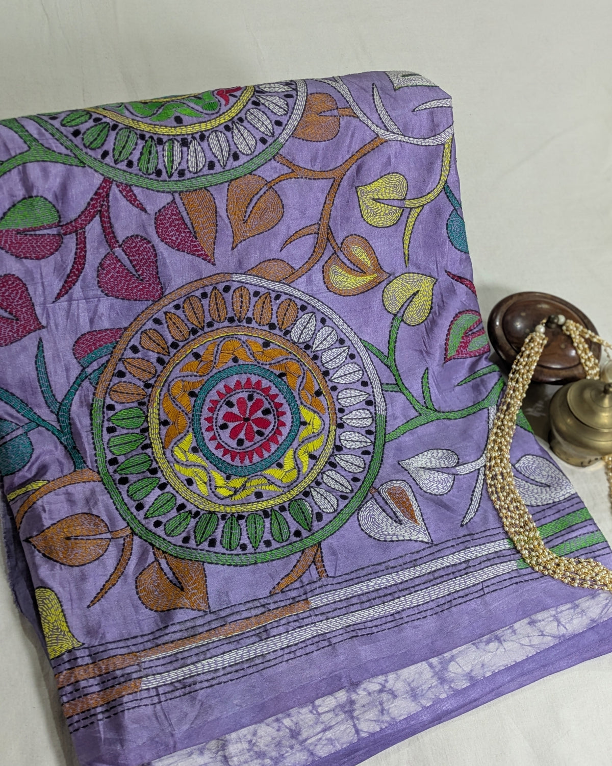 A folded purple fabric sari with intricate multicolor floral and mandala embroidery, shown on a light neutral background with a brass and wood decorative object partially visible on the right.