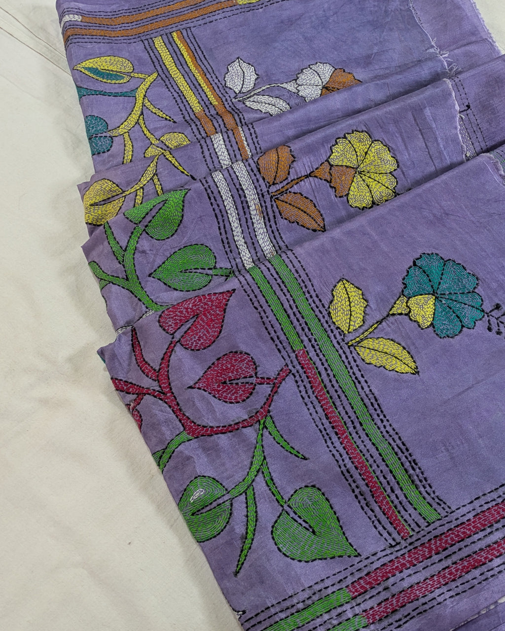 A folded lavender purple fabric, likely a cotton saree or textile, featuring colorful floral and vine embroidery in green, yellow, red, blue, and brown on a white background.