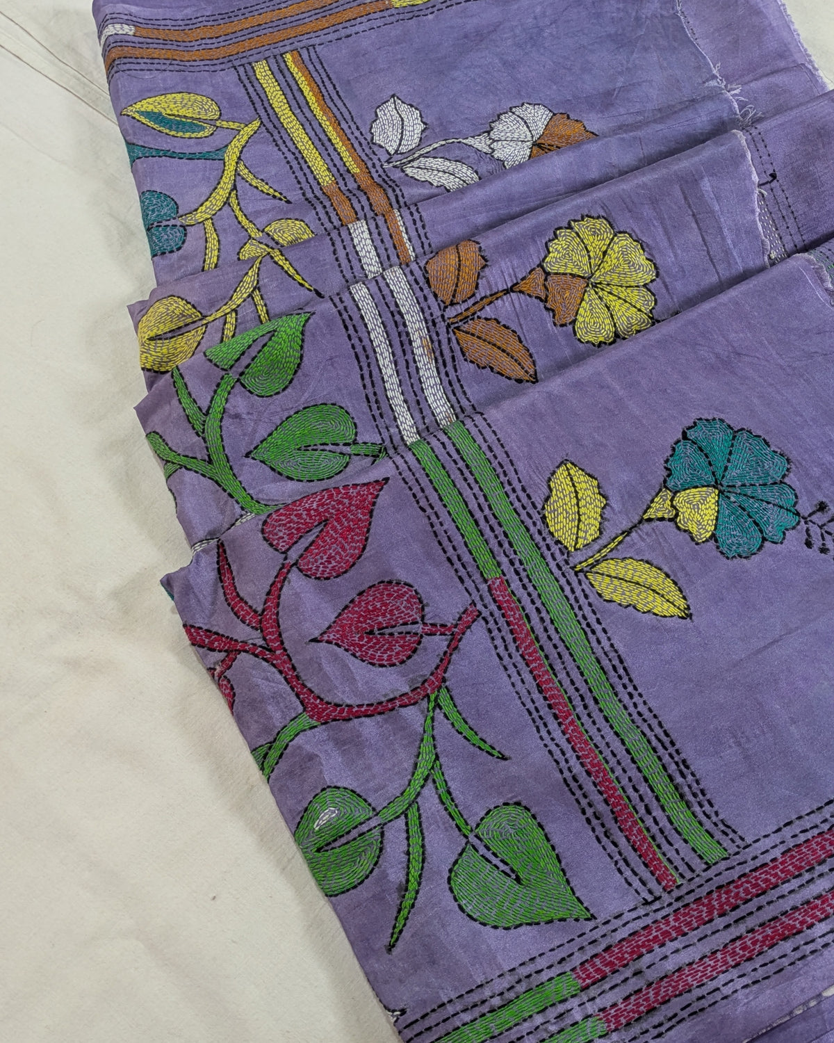 A folded lavender purple fabric, likely a cotton saree or textile, featuring colorful floral and vine embroidery in green, yellow, red, blue, and brown on a white background.