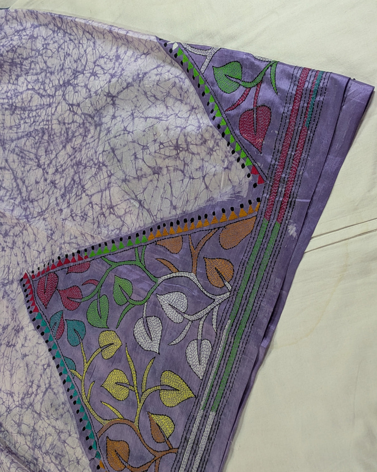 A close-up of a purple fabric textile, likely a saree or scarf, featuring a marbled pattern on one side and a decorative border with embroidered floral motifs in green, red, yellow, and orange on a lavender base.