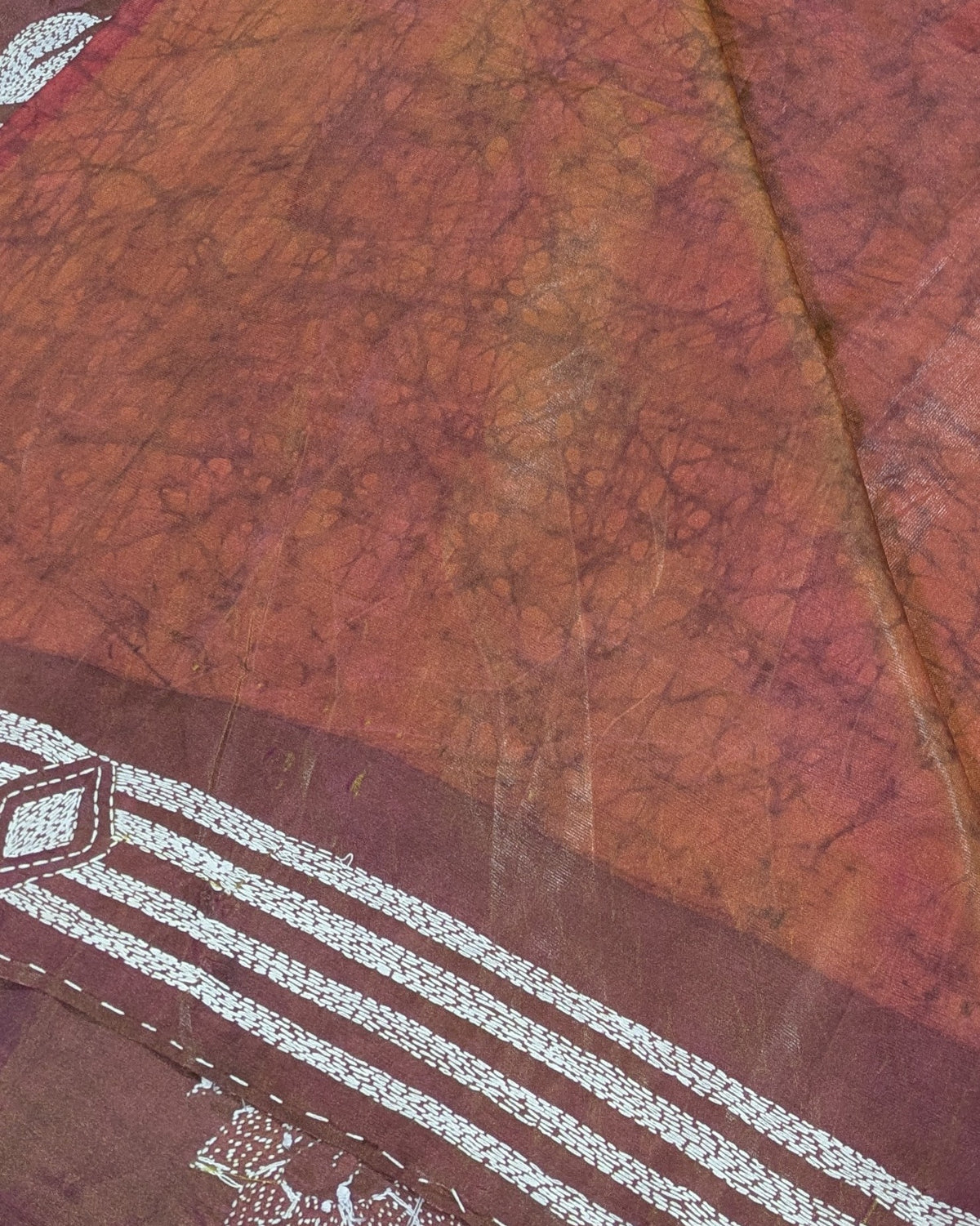 A close-up of a batik sari with a mottled orange, brown, and reddish-orange pattern, featuring a white and brown geometric striped border at the bottom.