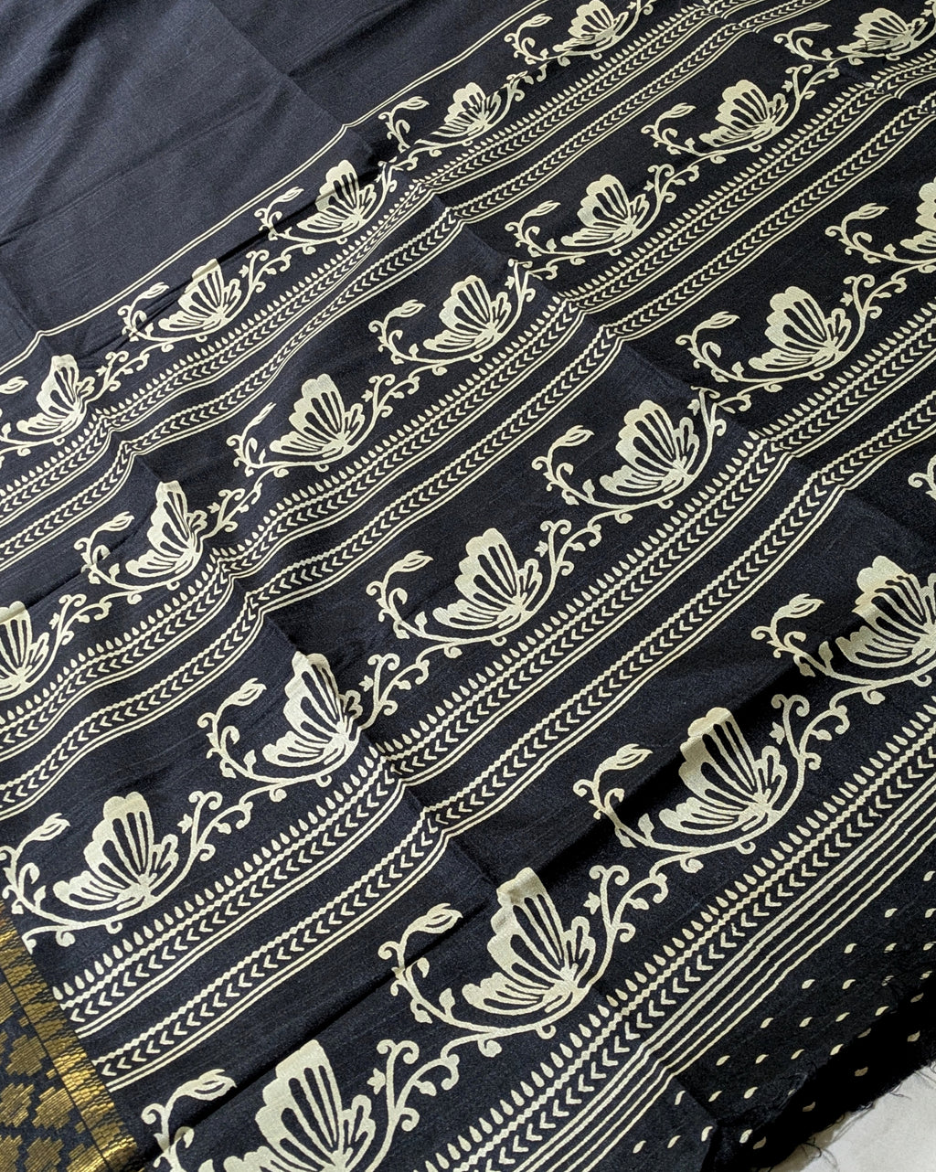 A close-up of a dark navy or black fabric with repeating cream floral and ornamental border prints, laid diagonally across the frame.