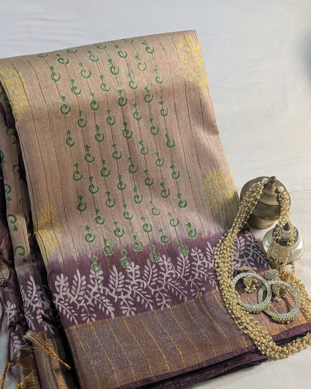 A folded traditional Indian saree displayed on a light fabric background, featuring a beige and brown body with green and yellow motifs, a purple border with white floral patterns, and styled with gold jewelry.