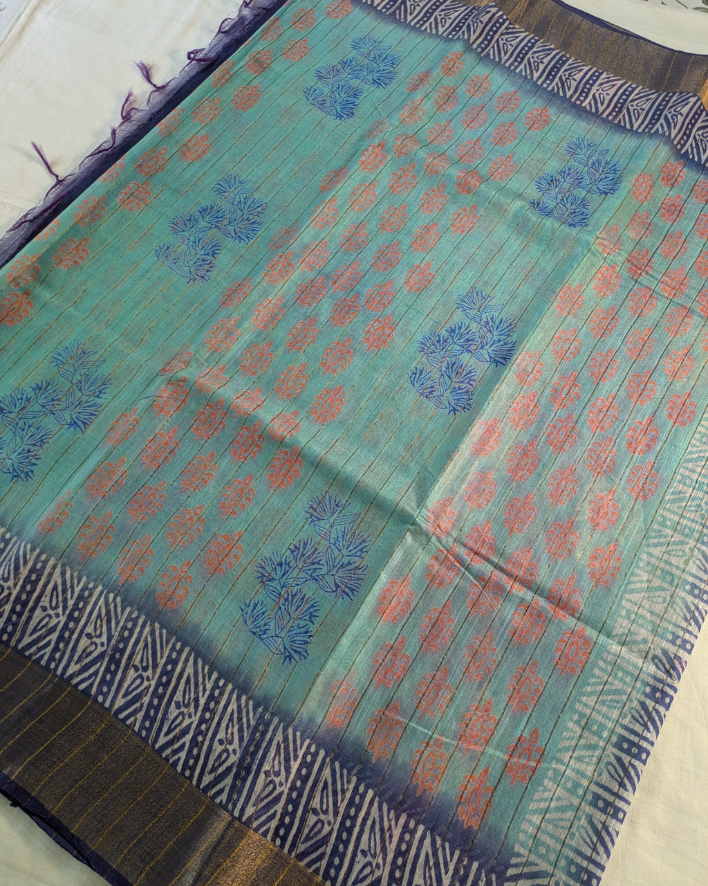 A flat-laid teal green saree with blue and pink floral block prints, featuring a dark navy and gold patterned border.