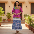 Woman in a pink top and blue floral skirt standing in front of a building with plants.
